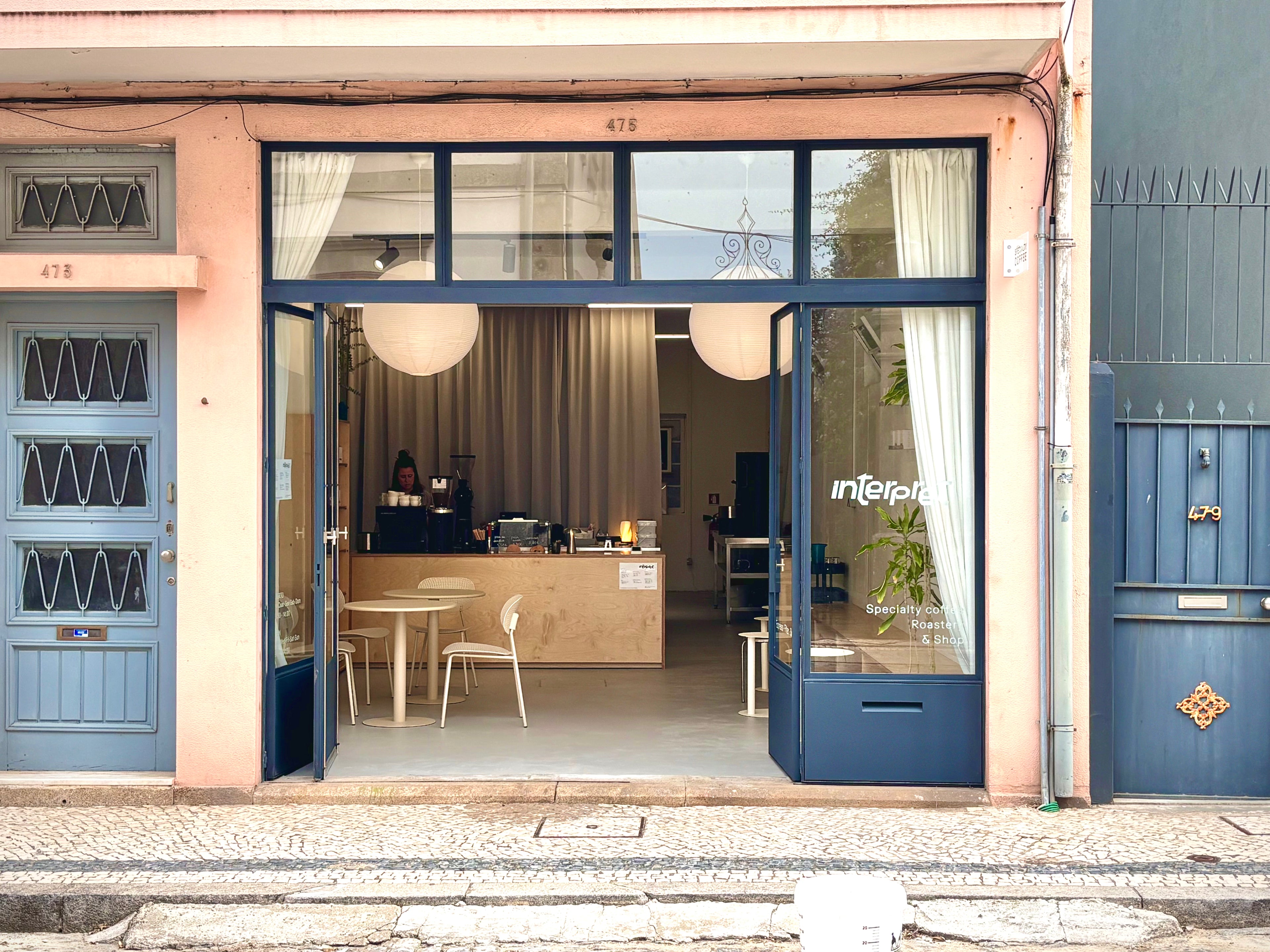 A storefront with a peach-pink facade and dark blue frames around large glass windows and doors. The cafe interior is visible through the open entrance, with tables, chairs, pendant lights, and a counter.