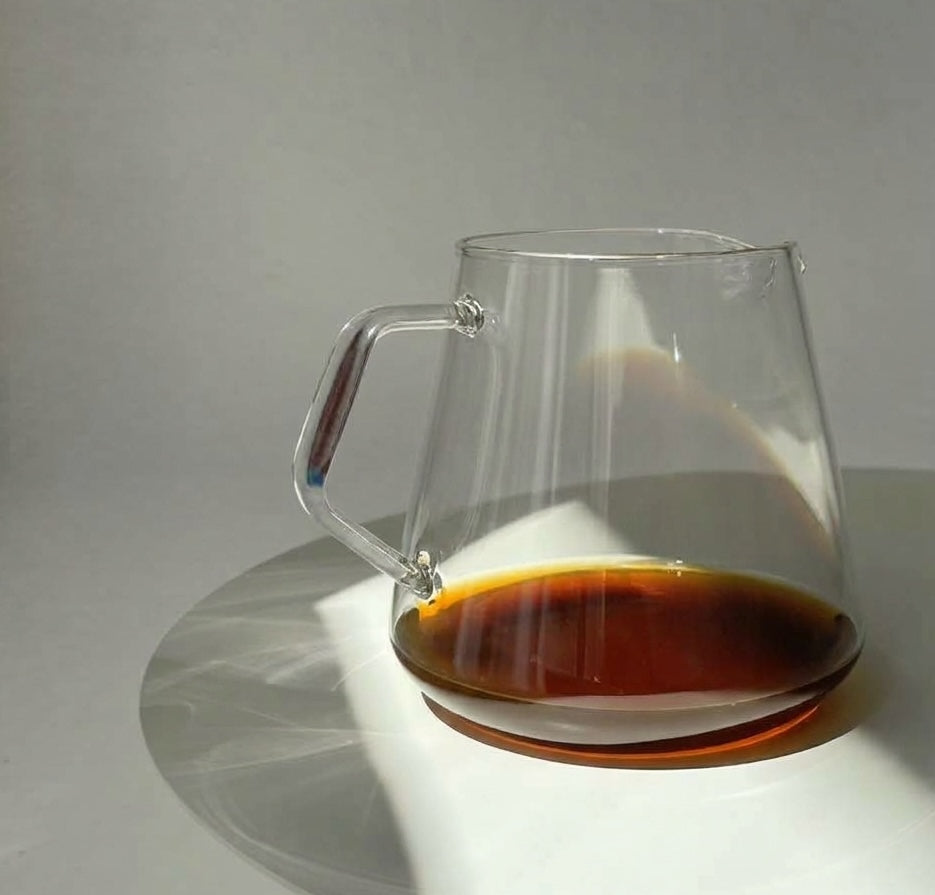 Clear glass pour-over coffee dripper with a handle, partially filled with dark coffee, sitting on a white surface with a soft shadow against a light gray background.